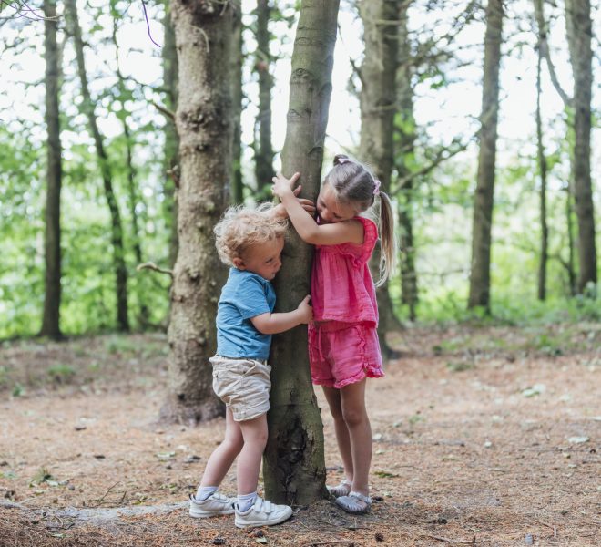 A brother and sister standing and hugging a tree in the woods.